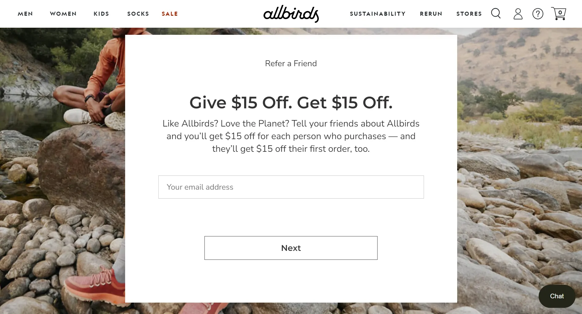 The Allbirds referral landing page features a background image of two people on a hike with stones in the background, symbolizing the brand's commitment to eco-friendliness. In the foreground, there is a white box with small and large font text that promotes their referral program, offering $15 off to both the referrer and the recipient. The page also includes a section to enter an email address and a call-to-action white button labeled "Next.”