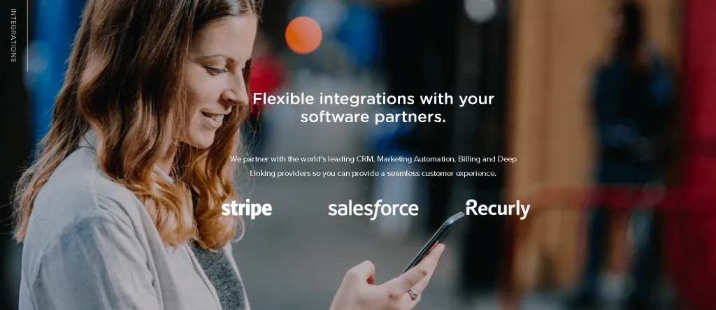 Smiling lady with brown curly hair wearing a ring on her middle finger, holding a smartphone showing Stripe, Salesforce and Recurly as SaaSquatch's software partners