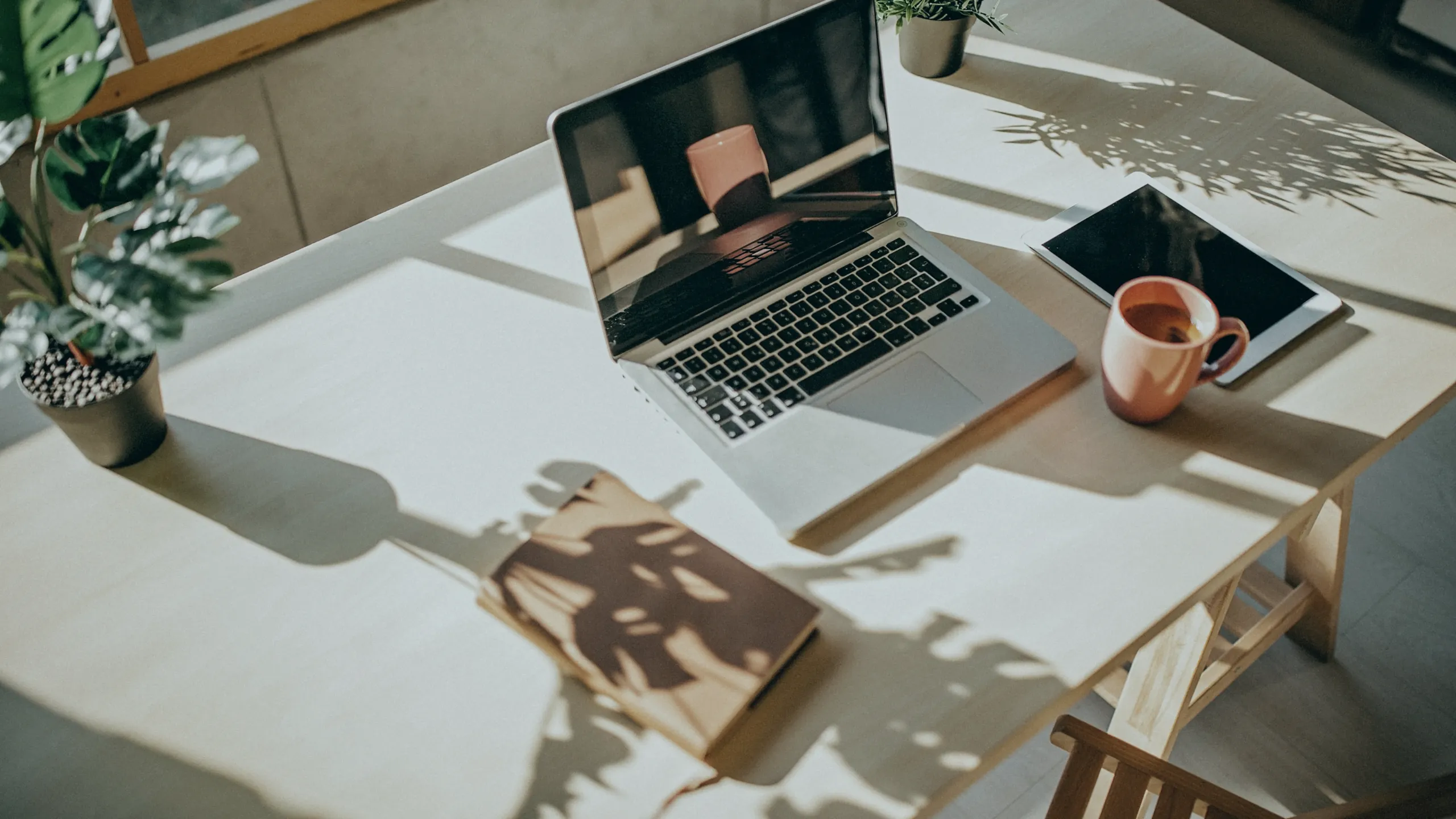 A bird's eye view of a wooden desk with a laptop and tablet. On the left corner, there is a small fake monstera plant and a beige notebook. To the right of the plant, there is a grey laptop. A pink coffee mug is next to the laptop and a tablet is next to the mug.