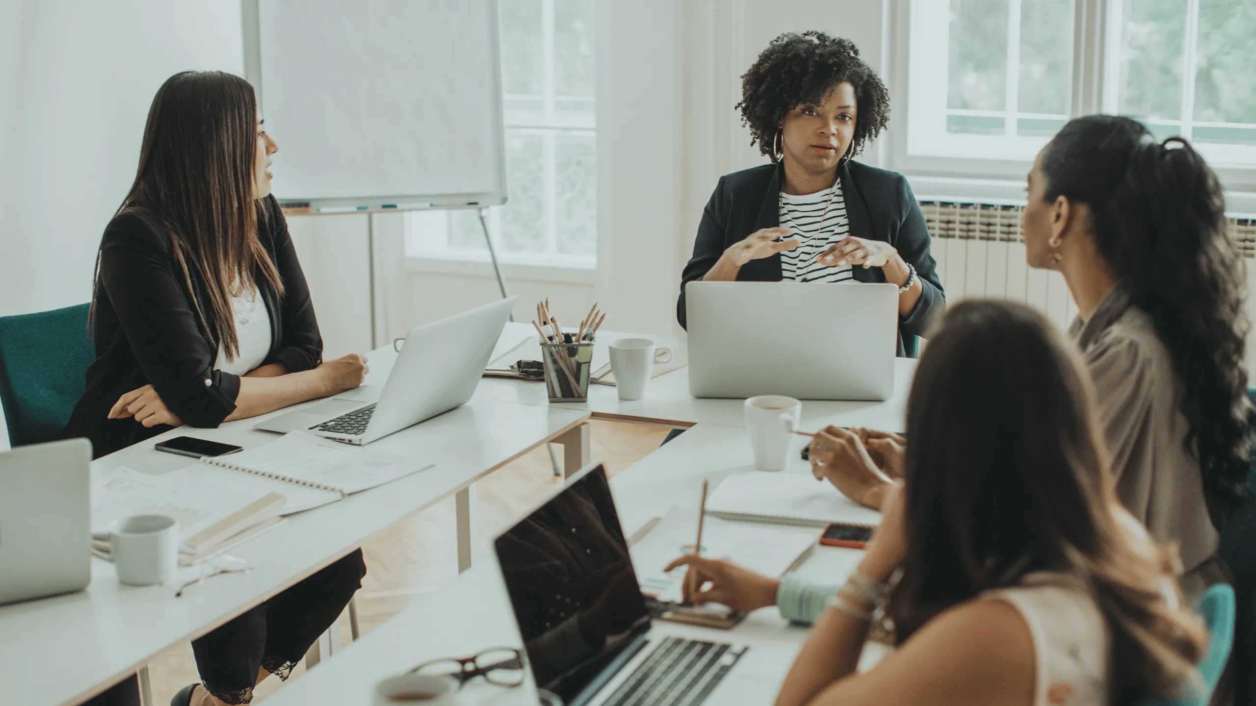 In the picture, four women are sitting around a table in an office environment. Three of them are looking at the woman sitting at the head of the table, who is gesturing with her hands while speaking. The women are all dressed in office attire and there are laptops, notebooks, pencils, and coffee cups on the table. The image represents a team evaluating the results of a loyalty program.