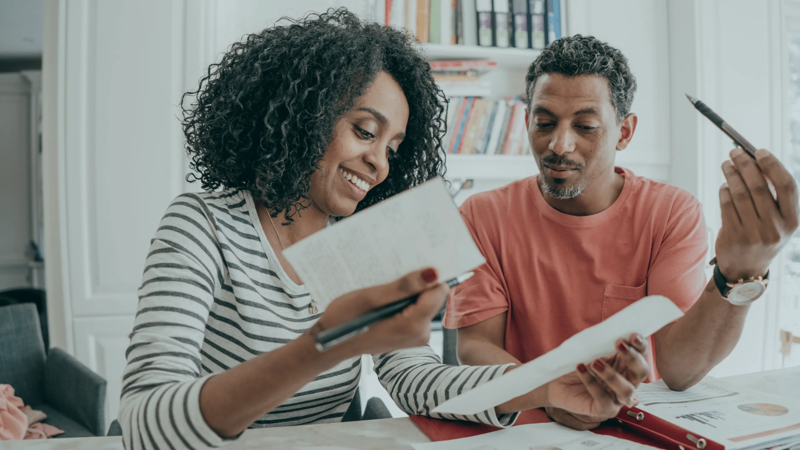 A man and a woman are sitting at a desk looking at papers and discussing a budget while books are visible in the background. The woman is smiling.