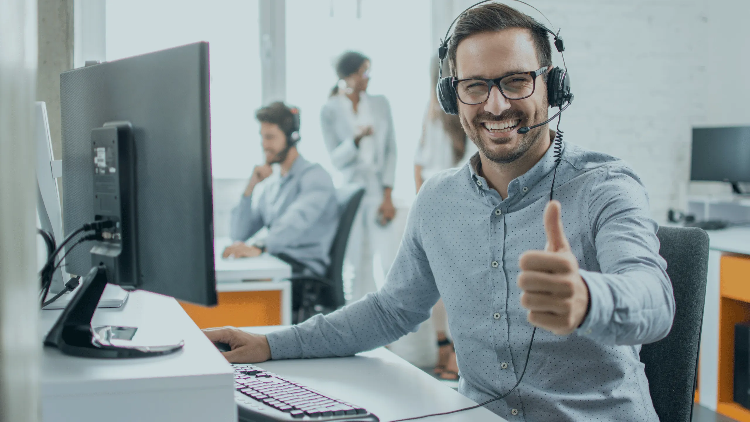 A man sitting at a desk with a computer and headphones, giving a thumbs up while smiling. Two other people can be seen in the background in an office environment.