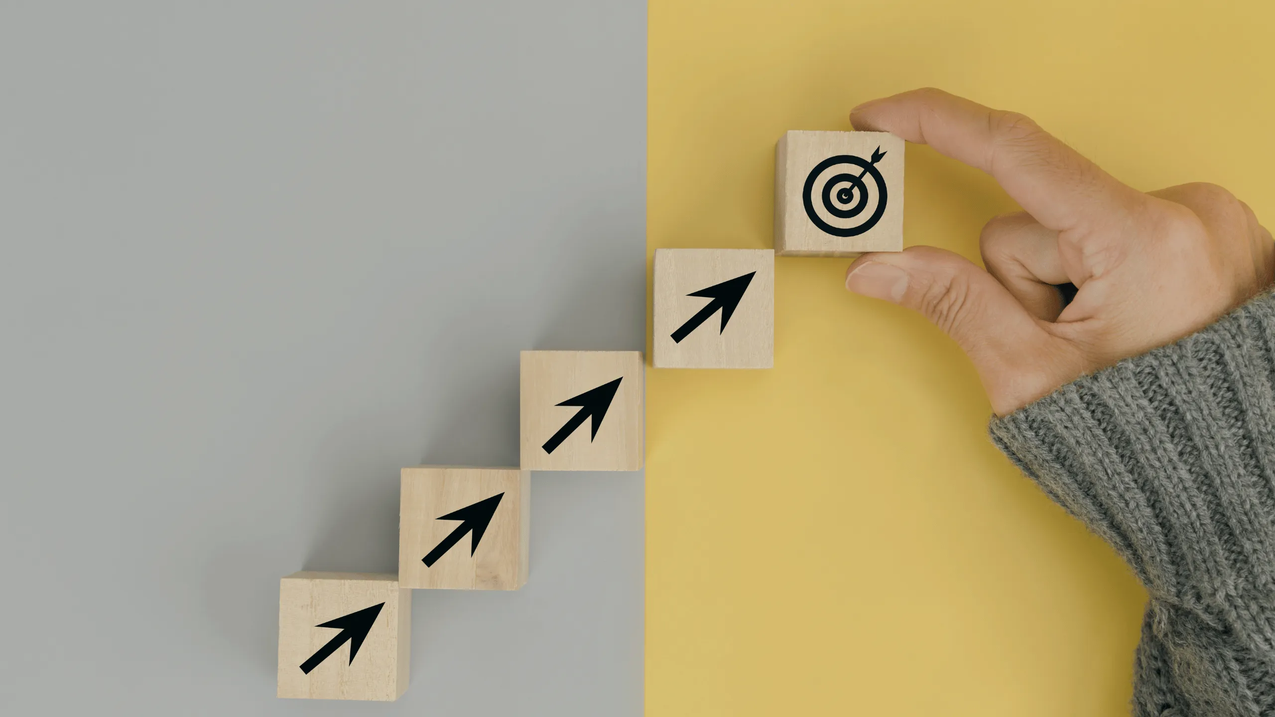 This image has a gray background on the left side and a yellow background on the right side. Four wooden boxes of equal size with arrow signs pointing towards a small wooden cell that has a target point on it, symbolizing achieving a goal through multiple process steps. A person wearing a grey sweater is holding the target box.