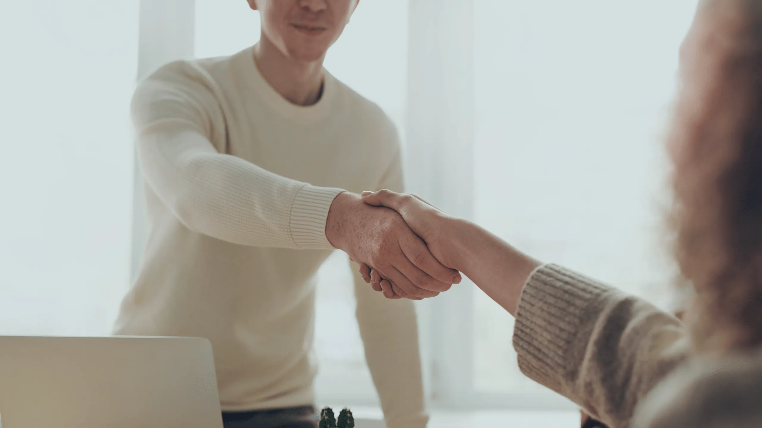 The image shows a handshake between two people. The first person is facing towards the viewer and is wearing a white sweater, while the second person is facing the first person and has long curly hair. The second person is wearing a gray sweater.