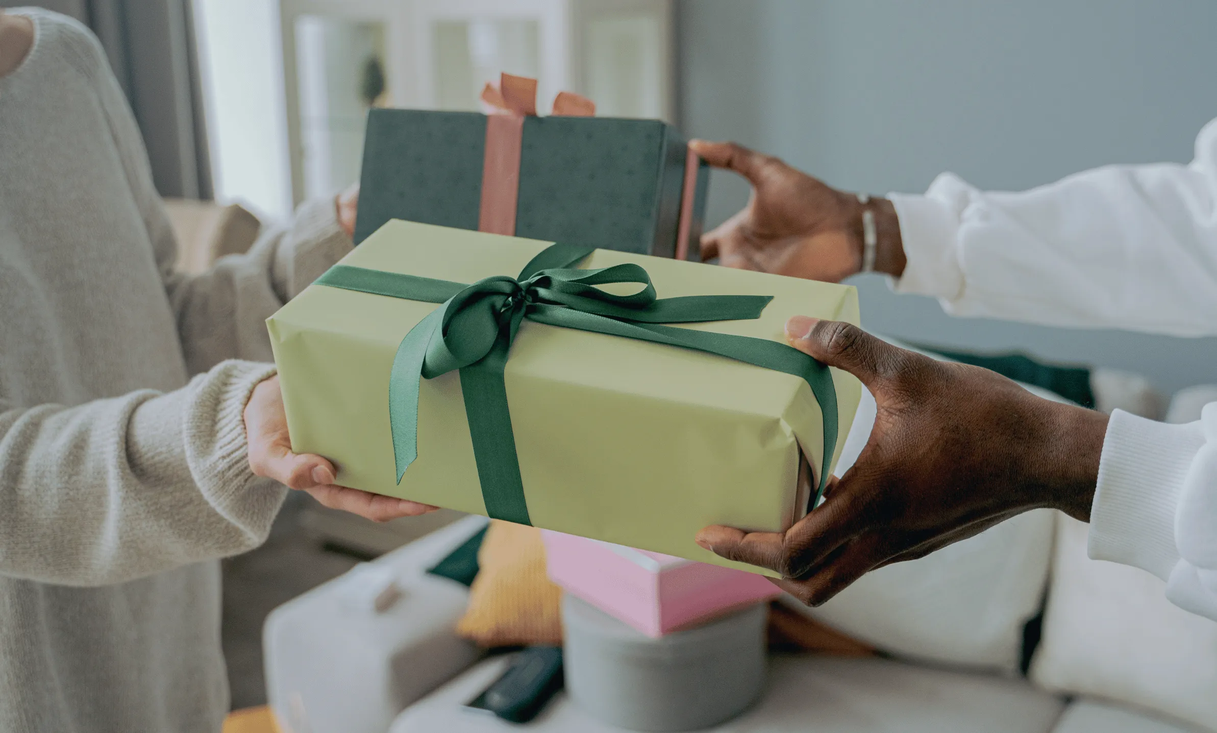 This photo shows two people exchanging gifts in a home setting. Only their hands and torsos are visible. The person on the left is wearing a beige sweater, while the person on the right is wearing a white sweater and has a darker skin tone. A blurred white couch can be seen in the background. The image is meant to symbolize the process of determining a rewards structure for a loyalty program.