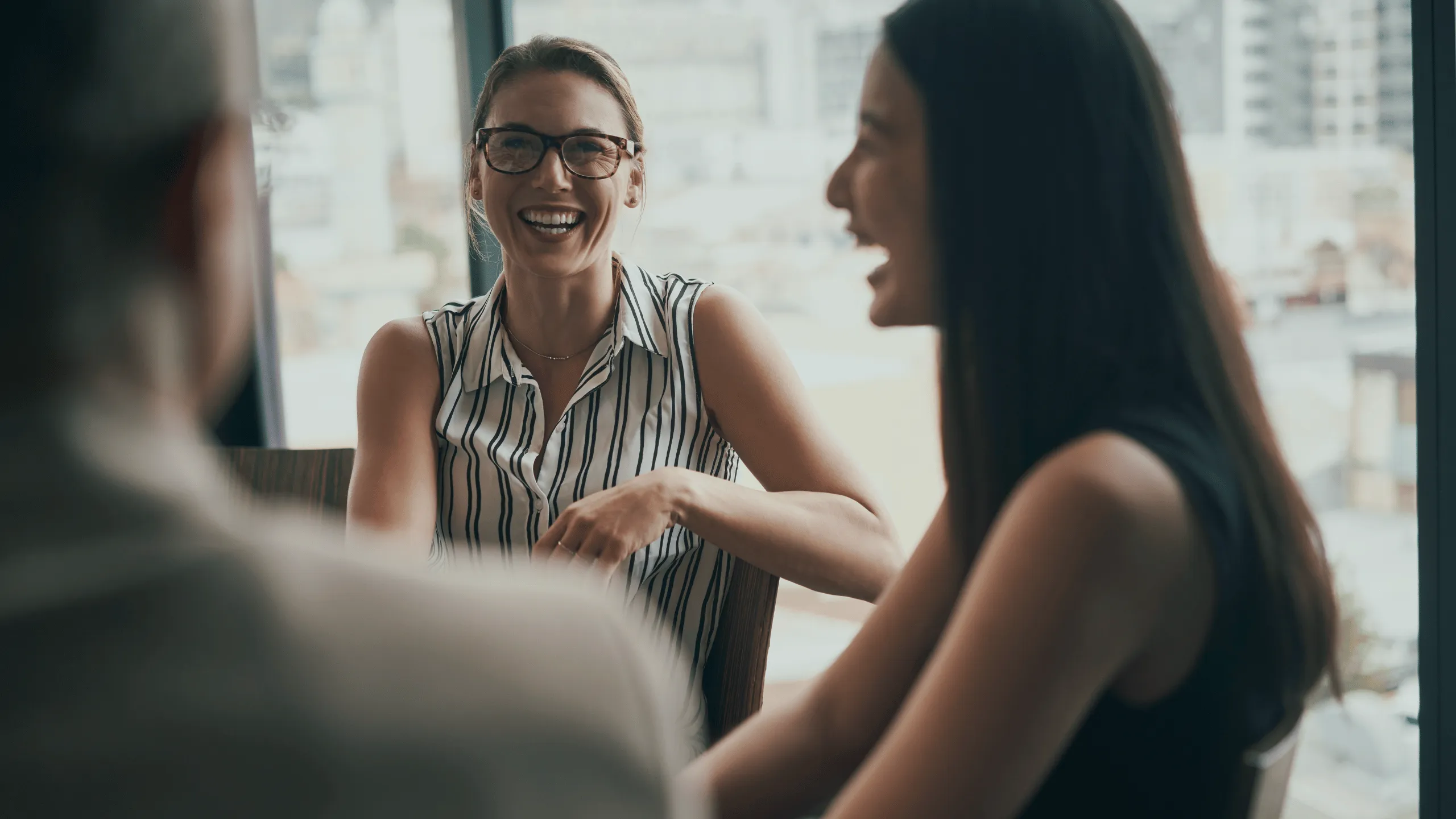  Three people in an office environment, two women and one man. One woman is facing towards the camera, while the other has her profile visible. The man is sitting with his back facing the camera. They are all laughing in a business professional manner.